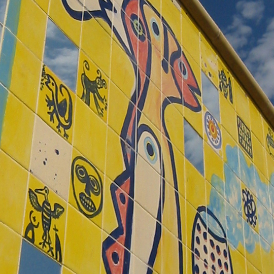 A mural made of yellow tiles with colourful hand panted designs against a blue sky on a sunny day